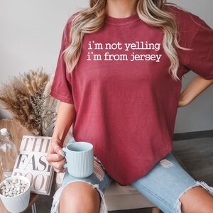 May include: A woman wearing a maroon t-shirt with the text "i'm not yelling i'm from jersey" in white. She is holding a light blue ceramic mug.