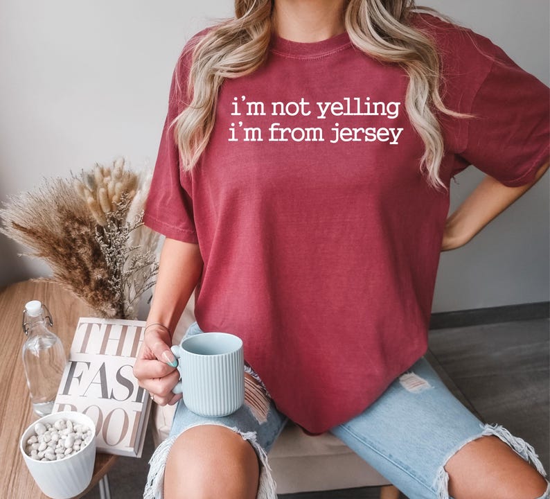 May include: A woman wearing a maroon t-shirt with the text "i'm not yelling i'm from jersey" in white. She is holding a light blue ceramic mug.