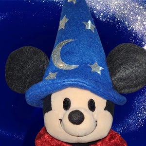 May include: A plush Mickey Mouse wearing a blue wizard hat with silver stars and a crescent moon. The hat is set against a dark blue background with a sparkling effect. Mickey has black ears and a red cape.
