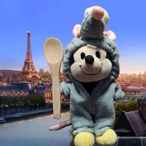 May include: A plush toy of a mouse wearing a gray hooded outfit and holding a wooden spoon. The mouse has a black nose and yellow feet. The background shows the Eiffel Tower and a cityscape.