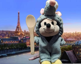 Könnte beinhalten: Ein Stofftier einer Maus in einem grauen Kapuzen-Outfit, die einen Holzlöffel hält. Die Maus hat eine schwarze Nase und gelbe Füße. Im Hintergrund sind der Eiffelturm und eine Stadtlandschaft zu sehen.