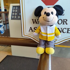 May include: A plush Mickey Mouse toy wearing a yellow and white vest with a stop sign in the background. The sign reads "STOP" in red letters. The sign also has the text "Wait for assistance".