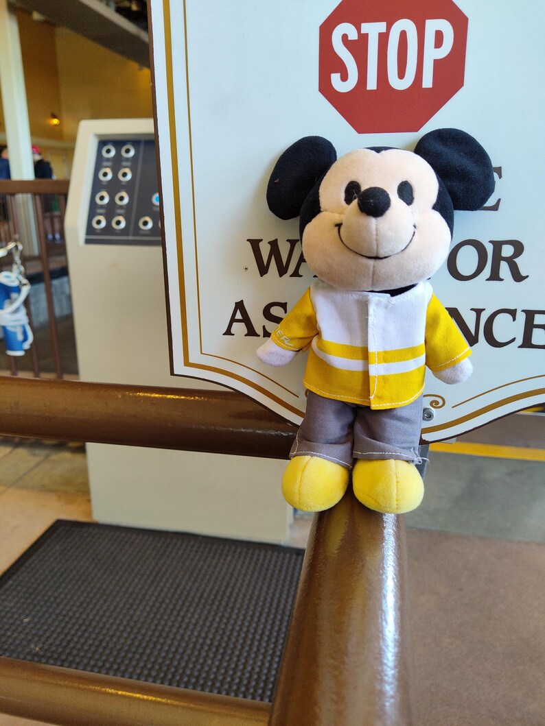 May include: A plush Mickey Mouse toy wearing a yellow and white vest with a stop sign in the background. The sign reads "STOP" in red letters. The sign also has the text "Wait for assistance".