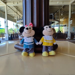 May include: Two plush toys of Mickey Mouse and Minnie Mouse wearing matching outfits. Mickey is wearing a yellow and white striped shirt with gray pants and yellow shoes. Minnie is wearing a blue and white striped shirt with gray pants and yellow shoes.