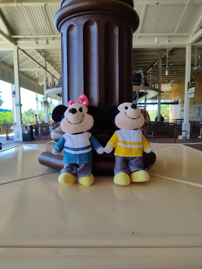 May include: Two plush toys of Mickey Mouse and Minnie Mouse wearing matching outfits. Mickey is wearing a yellow and white striped shirt with gray pants and yellow shoes. Minnie is wearing a blue and white striped shirt with gray pants and yellow shoes.