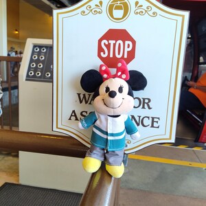 May include: A plush Mickey Mouse character wearing a green and white striped shirt and grey pants. The character is standing on a brown railing in front of a sign that says "STOP" and "Wait for Assistance".