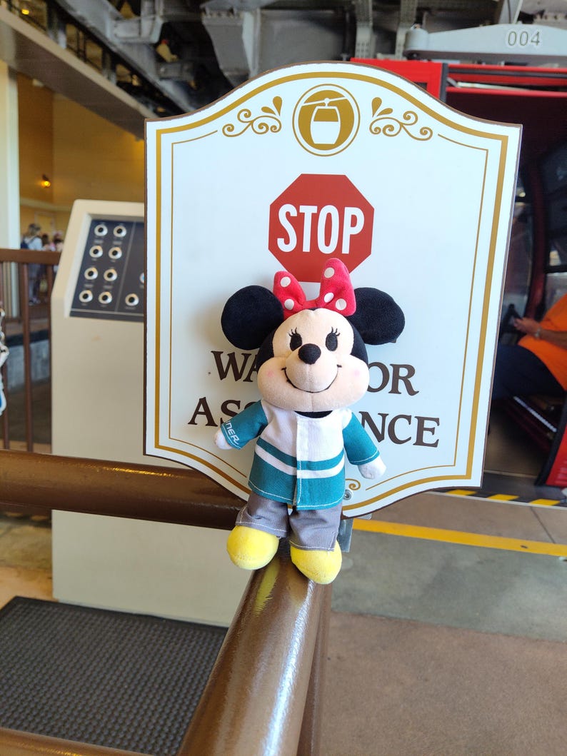 May include: A plush Mickey Mouse character wearing a green and white striped shirt and grey pants. The character is standing on a brown railing in front of a sign that says "STOP" and "Wait for Assistance".