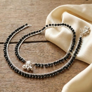 May include: A pair of anklets featuring a silver-toned chain adorned with small, faceted black beads. Each anklet includes a small silver-toned charm cluster consisting of three dangling bell-shaped elements attached to the main chain structure.