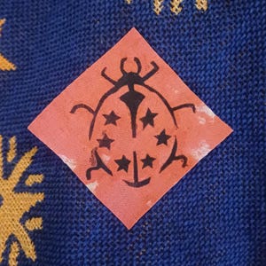 May include: A close-up of a square, coral-colored patch featuring a black ladybug design with star accents. The patch is set against a textured, dark blue fabric background with yellow and gold accents.
