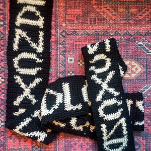 Peut inclure: Écharpe noire et blanche au crochet avec le mot "SOXODRO" en lettres blanches. L'écharpe est posée sur un tapis à motifs aux tons rouges, bleus et oranges. L'écharpe est faite à la main et semble douce et chaude.