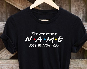 May include: Black t-shirt with white text that reads "The One Where NAME Goes To New York". The text is styled like the Friends logo with colorful dots.