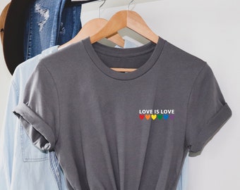 Könnte beinhalten: Ein graues T-Shirt mit dem Text "Love is Love" und einem Regenbogen aus Herzen auf der Vorderseite.
