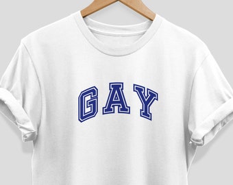 Op de afbeelding: Wit T-shirt met het woord "GAY" in blauwe, gebogen, collegiale letters. Het shirt wordt op een houten hanger gepresenteerd tegen een neutrale achtergrond. De mouwen zijn opgerold.