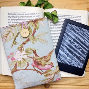 Può includere: Una custodia per libro blu chiaro con un motivo floreale con uccelli rosa e verdi. La custodia ha una chiusura a bottone ed è progettata per adattarsi a un lettore elettronico Kindle. Il Kindle è visibile nell'immagine, che mostra un'immagine in bianco e nero di uno strumento musicale.