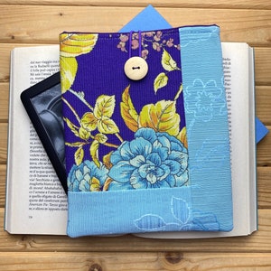 Könnte beinhalten: Ein Buchcover aus lila und blauem Stoff mit einem Blumenmuster. Das Cover hat einen Knopfverschluss und ein hellblaues Band als Lesezeichen. Das Cover liegt auf einem Buch.