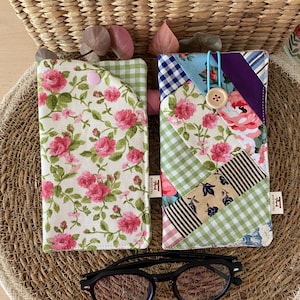 Könnte beinhalten: Zwei Stoff-Brillenetuis mit Blumen- und Patchwork-Designs, zusammen mit einer Brille mit schwarzem Rahmen. Ein Etui zeigt rosa Rosen auf weißem Grund, das andere ein Patchwork aus Mustern. Die Etuis liegen auf einer gewebten Matte.