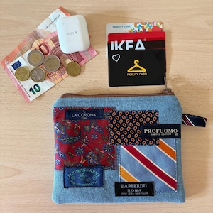 Puede incluir: Un estuche de cremallera en tela vaquera azul claro con parches de tela en motivos florales rojos, lunares azules y rayas. Las etiquetas incluyen La Corona, Profuomo, Thomas & Kent y Barberini Roma. Accesorios: estuche de auriculares blanco, billetes de euro, monedas y tarjetas de fidelidad.