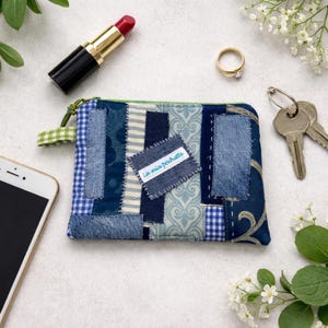 Peut inclure: Une petite pochette en denim patchwork avec une fermeture éclair verte et une petite étiquette avec l'inscription "La mia pochette". Un rouge à lèvres rouge, une bague en or, des clés et un smartphone sont également visibles.