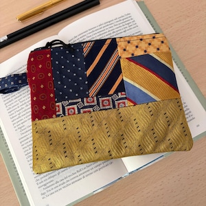 May include: A rectangular zippered pouch constructed from a patchwork of various silk neckties in shades of red, navy blue, and gold, featuring a prominent bottom panel of textured gold fabric with a repeating wave pattern and a black zipper closure.