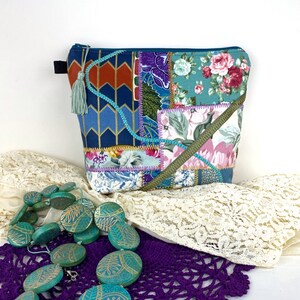 Puede incluir: Una bolsa de cremallera de tela patchwork con un fondo azul verdoso y patrones florales en tonos de rosa, morado y verde. La bolsa tiene un borla azul verdoso.