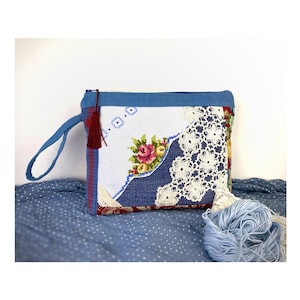 Può includere: Una pochette blu con cerniera, manico in tessuto e nappina. La pochette è realizzata in tessuto patchwork con un motivo floreale, pizzo e denim blu. La pochette è appoggiata su uno sfondo in tessuto blu.