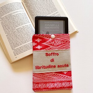 Può includere: Una custodia in tessuto rosso e bianco con un motivo a fantasia e una patta beige. La patta presenta il testo "Soffro di libritudine acuta" ricamato in rosso. La custodia è progettata per contenere un e-reader.