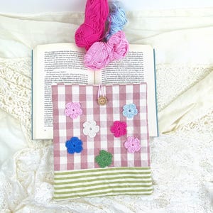 May include: A pink and white gingham book cover with a green and white striped bottom. The cover has a button closure and is decorated with colourful crocheted flowers.