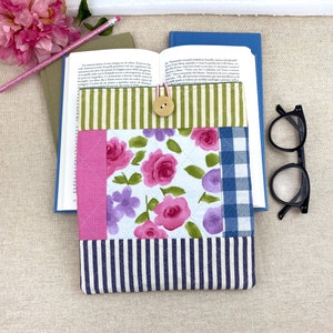 Può includere: Una custodia per libri trapuntata con motivi floreali e a righe. La custodia presenta rose rosa, fiori viola e tessuto a quadretti blu. Un bottone di legno e un elastico rosa fissano la custodia. Sono visibili anche una matita e degli occhiali.