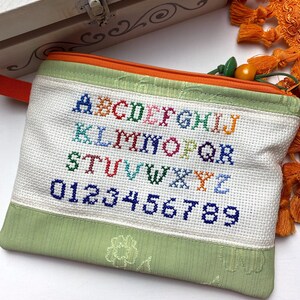 Può includere: Una pochette con cerniera verde e arancione con sfondo bianco. La pochette presenta un ricamo a punto croce dell'alfabeto e dei numeri in vari colori.