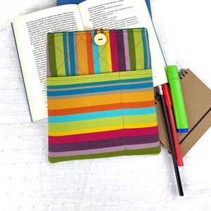 May include: A colourful striped fabric book cover with a button closure and a pocket. The cover is lying on a white surface with a book, a notebook and pens.