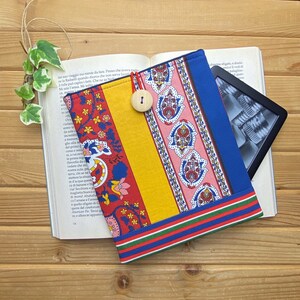Könnte beinhalten: Ein bunter Stoffbuchumschlag mit einem blauen, gelben, roten und pinkfarbenen Muster. Der Umschlag hat einen Knopfverschluss und ist für einen E-Reader konzipiert.