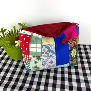 Peut inclure: Une pochette zippée en patchwork avec une doublure rouge et un pompon. La pochette est faite de différents tissus avec divers motifs, notamment des pois, du tartan, des fleurs et du paisley.