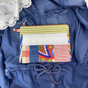 Può includere: Una pochette con cerniera in tessuto patchwork con un bordo in pizzo bianco, motivi rossi, blu e verdi e una parte superiore a righe verde chiaro e bianco. La pochette è appoggiata su una camicia di jeans blu.