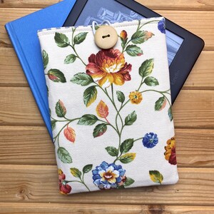 Può includere: Una custodia per Kindle in tessuto bianco con motivo floreale e chiusura a bottone in legno. La custodia è mostrata con un libro blu e un e-reader Kindle nero.