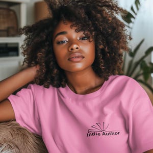 Könnte beinhalten: Ein pinkfarbenes T-Shirt mit dem Text "Indie Author" und einer Buchgrafik. Das T-Shirt wird von einer Person mit dunklem, lockigem Haar getragen. Das Bild ist eine Nahaufnahme.