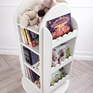 May include: White, rotating bookshelf with scalloped edges, filled with children's books and stuffed animals. The bookshelf has three shelves and a circular base. Books include "The Midnight Fair" and other colorful titles.