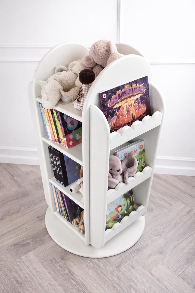 May include: White, rotating bookshelf with scalloped edges, filled with children's books and stuffed animals. The bookshelf has three shelves and a circular base. Books include "The Midnight Fair" and other colorful titles.