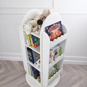 May include: A white, rotating bookshelf with scalloped edges, filled with colorful children's books and stuffed animals. The bookshelf has three shelves and a circular base. The books are of various sizes and colors. The overall theme is children's decor.