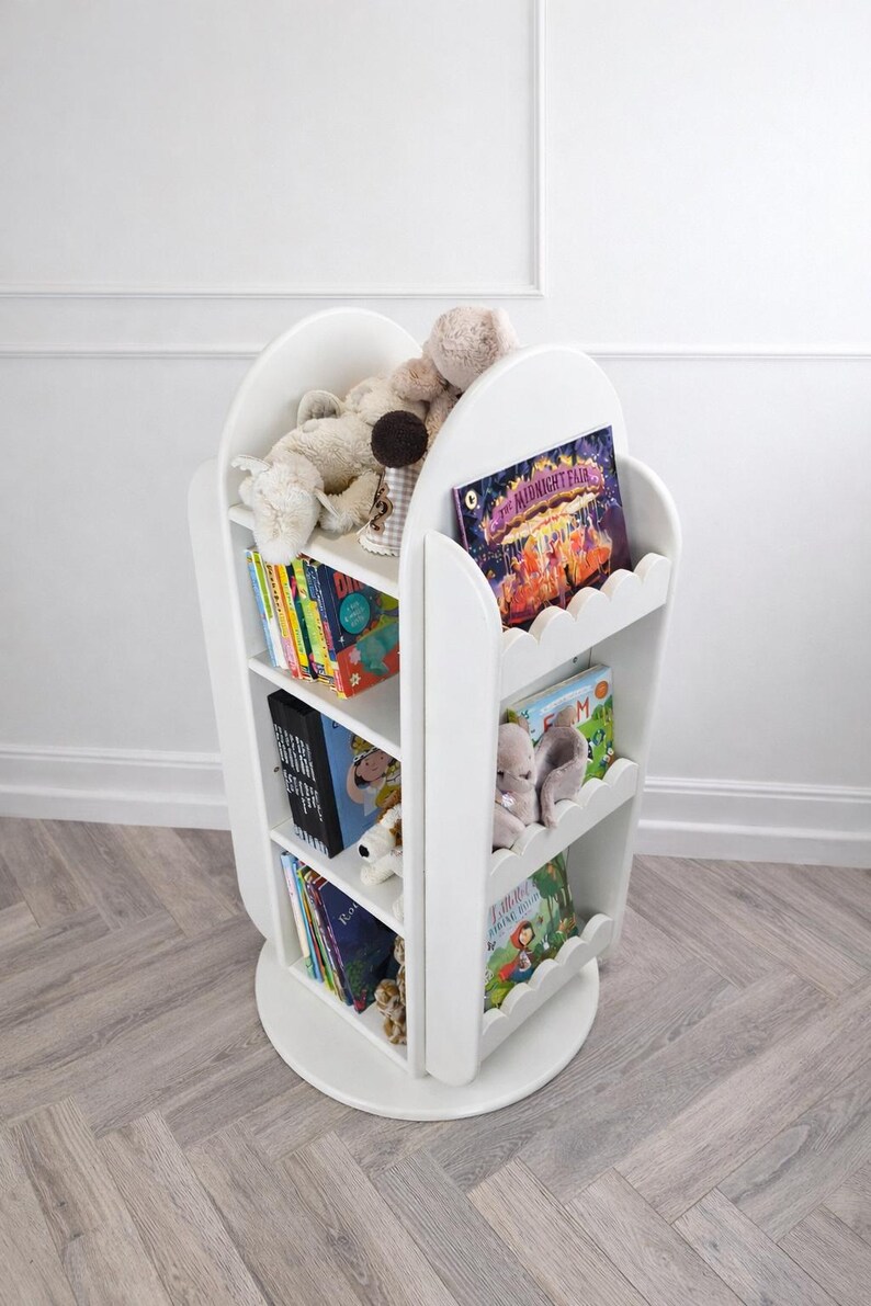 May include: A white, rotating bookshelf with scalloped edges, filled with colorful children's books and stuffed animals. The bookshelf has three shelves and a circular base. The books are of various sizes and colors. The overall theme is children's decor.