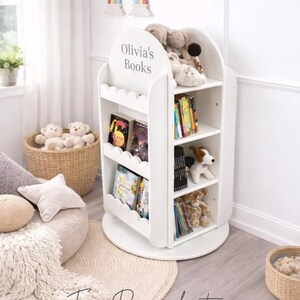 May include: White, three-tiered bookshelf with scalloped edges, labeled "Olivia's Books." The shelves hold books and stuffed animals. A rainbow decoration hangs above the bookshelf. A woven basket and a round rug are in the foreground.