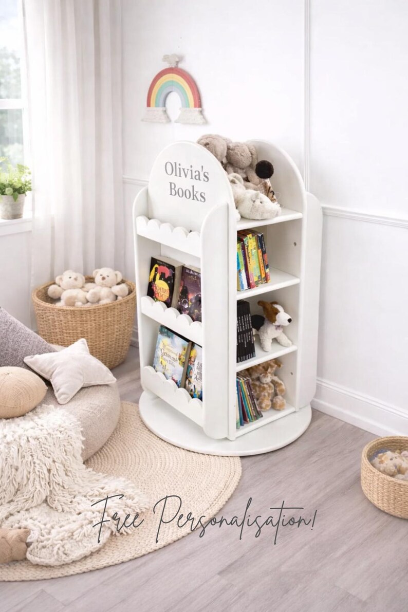 May include: White, three-tiered bookshelf with scalloped edges, labeled "Olivia's Books." The shelves hold books and stuffed animals. A rainbow decoration hangs above the bookshelf. A woven basket and a round rug are in the foreground.