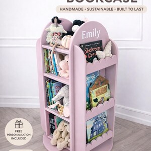 May include: A pink, personalized children's bookcase with the name "Emily" on top. The bookcase is round with scalloped edges and multiple shelves filled with books and stuffed animals. The text "HANDMADE &bull; SUSTAINABLE &bull; BUILT TO LAST" is visible.