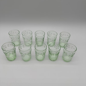 May include: A set of ten light green glass tumblers arranged in two rows. Each tumbler features a cylindrical shape with a slightly flared rim and a repeating etched pattern of small circular dots encircling the upper portion of the glass.