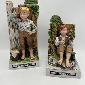 May include: Two ceramic figurines depicting characters from the Tom Sawyer novel. One figure is Tom Sawyer, leaning against a wooden fence. The other is Huck Finn, seated on a rock. Both figurines have green foliage accents and are on rectangular bases.