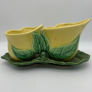 May include: A yellow ceramic planter with green leaf details. The planter is divided into two sections and has a matching green leaf-shaped base.