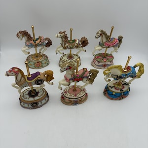 May include: A collection of seven miniature carousel horse figurines. Each horse is a different color and design, with gold poles and decorative bases. The horses have detailed manes, tails, and saddles, and are set against a white background.