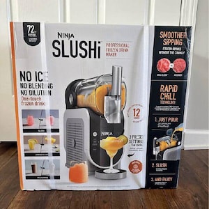 May include: A Ninja Slushi professional frozen drink maker in a box. The box is white and black with the words "Ninja Slushi" in large black letters. The box also shows images of the machine and frozen drinks. The machine is black and silver.