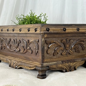Może przedstawiać: Ciemnobrązowa drewniana skrzynia z rzeźbionymi wzorami kwiatowymi. Prostokątna skrzynia ma płaską górę i jest wsparta na czterech ozdobnych nóżkach. Małe, ciemne metalowe ćwieki obramowują górną krawędź. Na górze znajduje się zieleń.