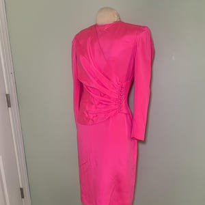 May include: A bright pink dress with long sleeves and a knee-length skirt. The dress features a draped design on the bodice and a row of buttons down the side. The dress is displayed on a wooden dress form.