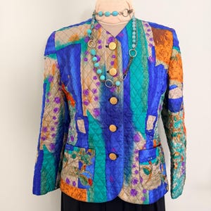 May include: A quilted jacket featuring a vibrant abstract pattern in shades of blue, teal, orange, and purple with gold-tone buttons. A multi-strand necklace with turquoise-colored beads and silver-tone metal links is draped over the collar.